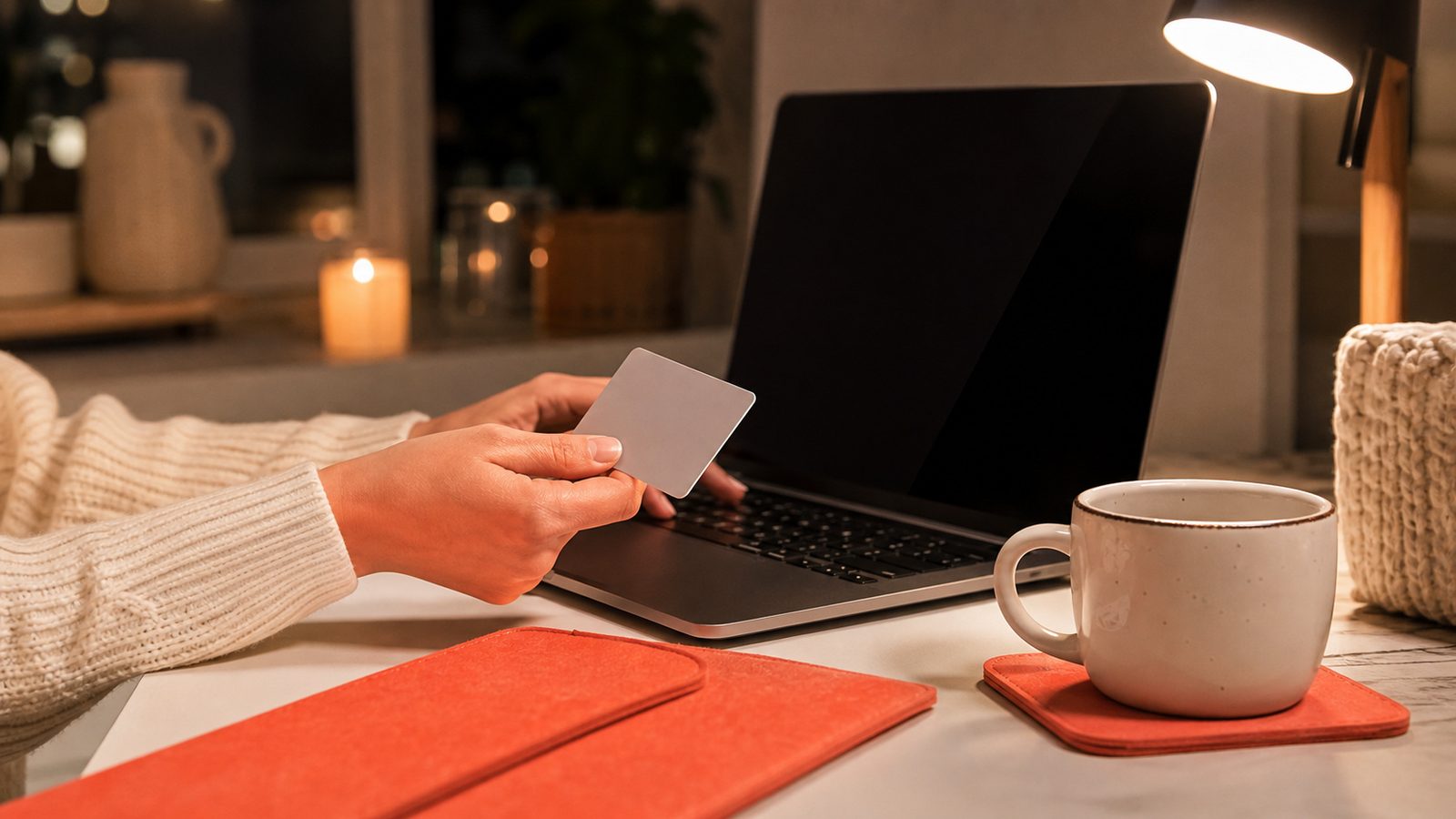 Dłonie z kartą płatniczą przy laptopie, obok kubek na koralowej podkładce